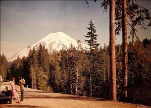 A white man and woman in fancy clothes stand on the side of a dirt road taking pictures of a snow capped mountain